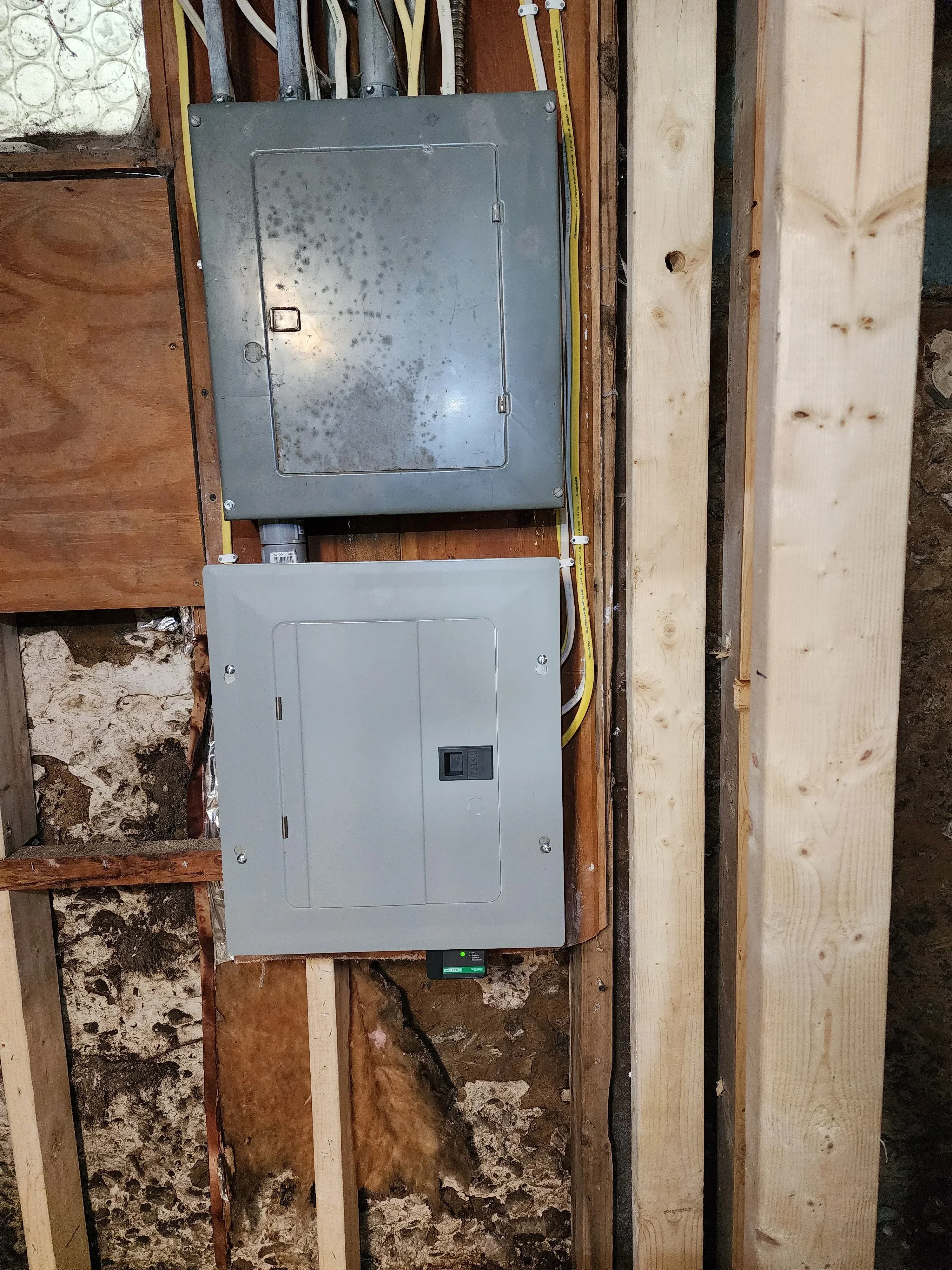 Two electrical breaker panels mounted on wood framing with yellow and white conduit wiring